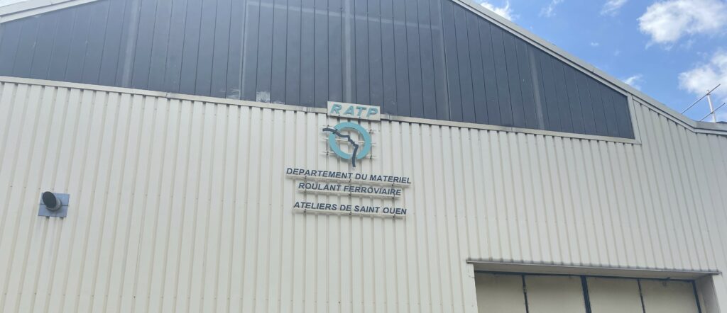 Photographie de la face d'un hangar de la RATP. Le hangar est très grand. Le logo de la RATP est accroché sur la face avant et on peut lire distinctement : Département du matériel roulant ferroviaire, ateliers de Saint-Ouen.
